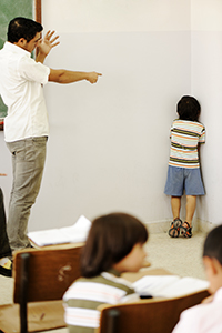 Imagem de uma sala de aula com professor apontando o dedo em direção a um aluno de castigo no canto da parede.